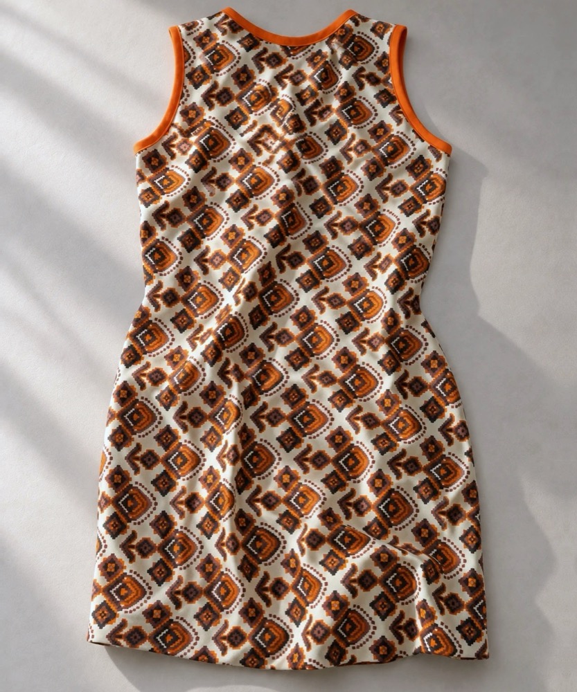 The older piece laid flat in window light: a sleeveless shift dress in a brown and orange print with orange trim at the neck and arms.