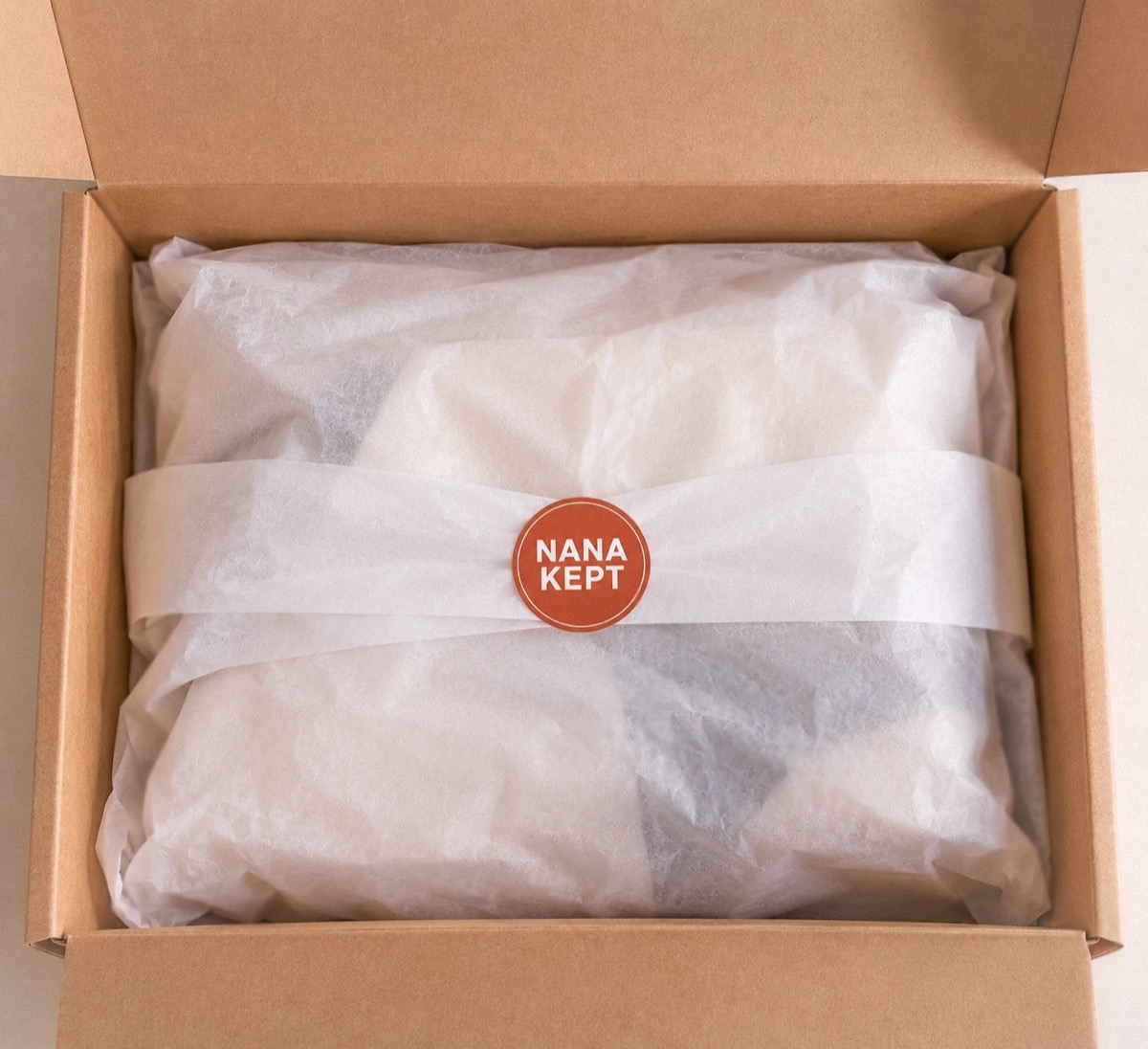 A kraft shipping box open on a table, the pieces wrapped in white tissue and closed with a round Nana Kept sticker.